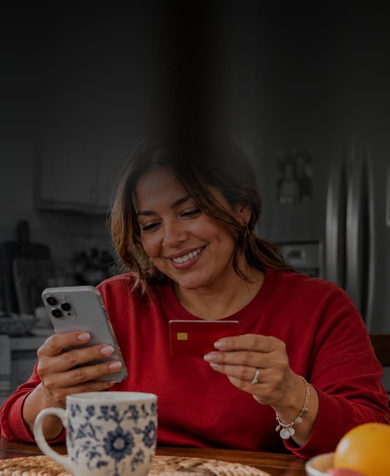 Una mujer en la mesa de su cocina pagando su seguro desde el celular con una tarjeta